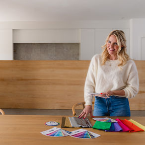 Smiling woman in white sweater holds color swatches at a wooden table. Colorful fabric samples and charts are laid out. Bright, modern room.