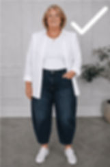 Smiling older woman in white shirt, dark jeans, and sneakers stands indoors against a gray wall with a large white checkmark.