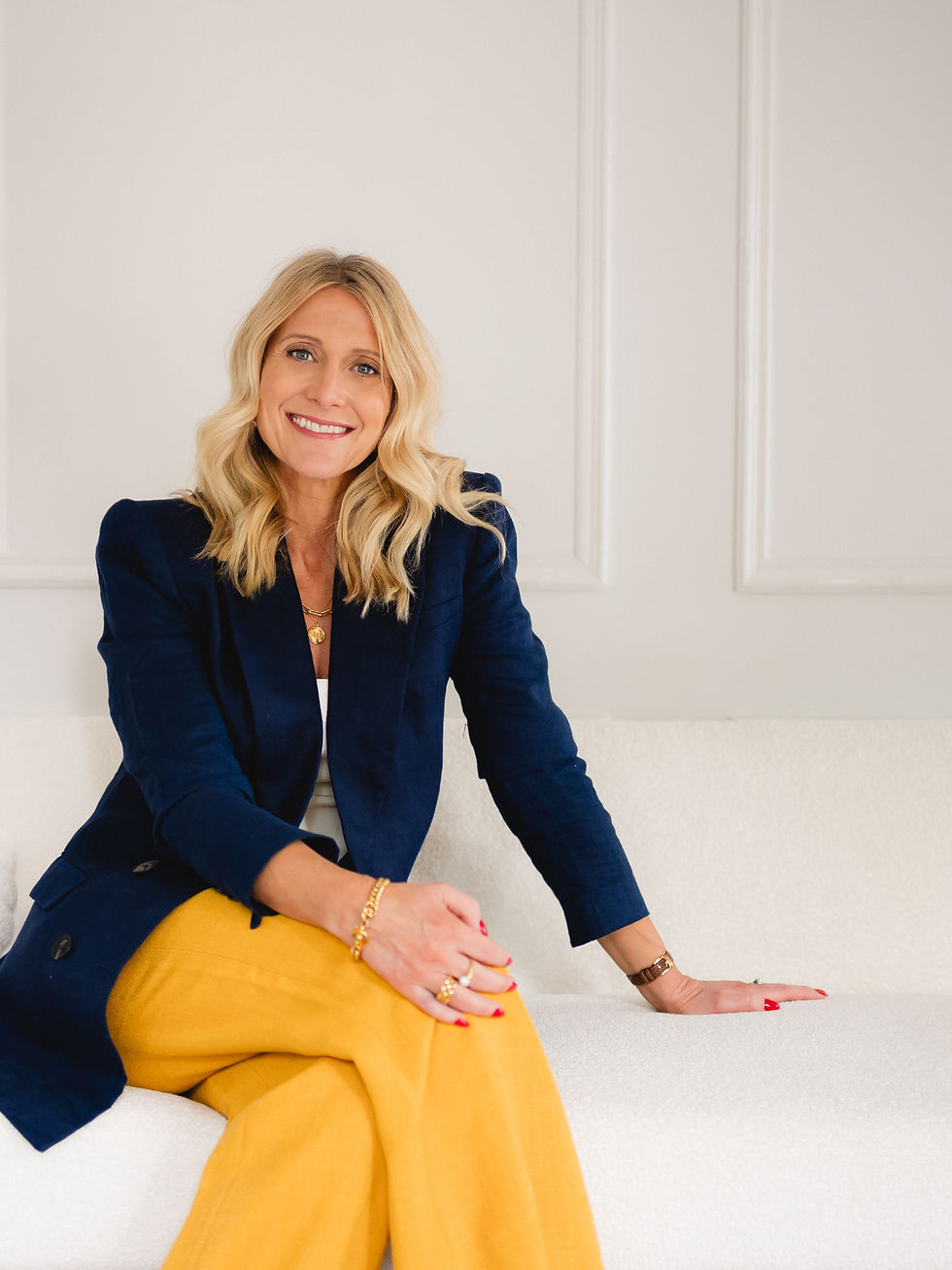 Melissa sits on a white sofa facing the camera and smiling. She is wearing a navy blue blazer and yellow trousers