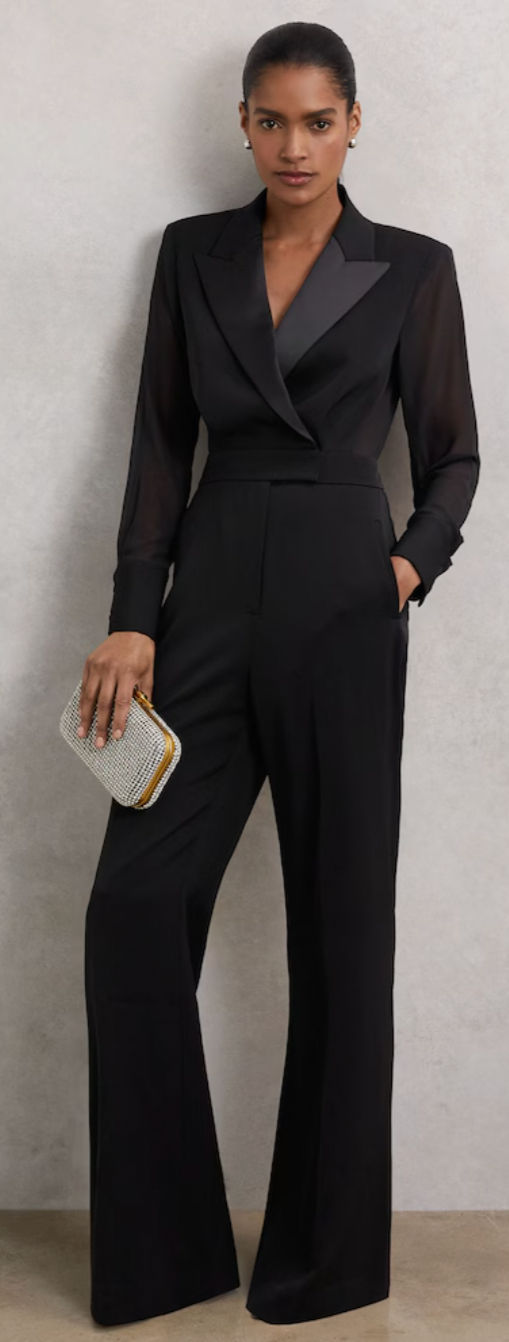 Woman in black pantsuit with sheer sleeves holds a silver clutch. Neutral background, confident expression, elegant and poised.