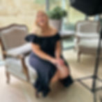 Jess sits on a chair smiling at the camera wearing a black off the shoulder dress