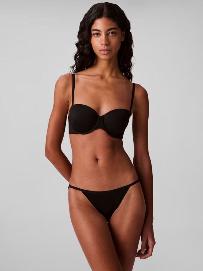Person in a black bikini stands confidently against a plain gray background. Their expression is neutral, and the lighting is soft.