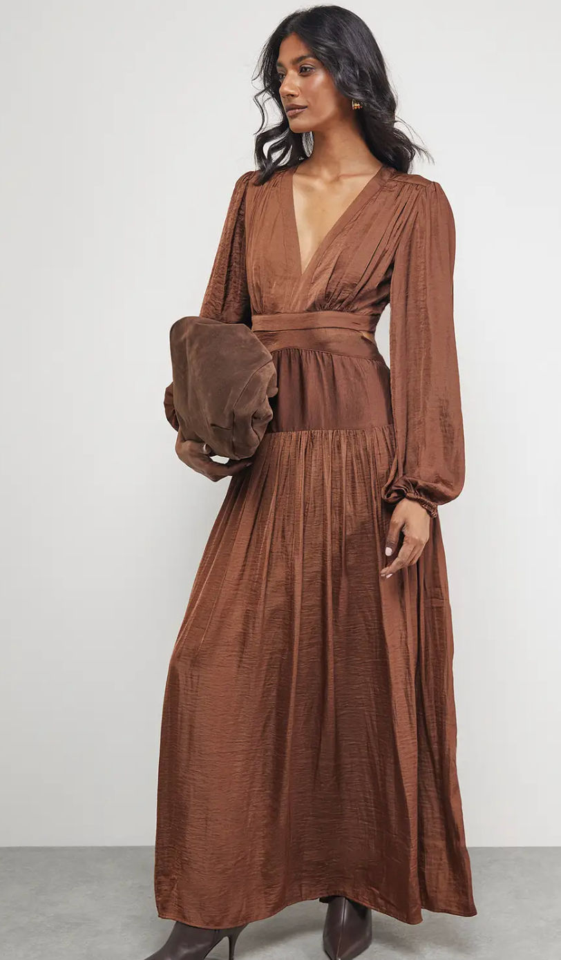 Woman in a brown long-sleeve dress holding a matching brown clutch. She's standing against a light gray background, exuding elegance.