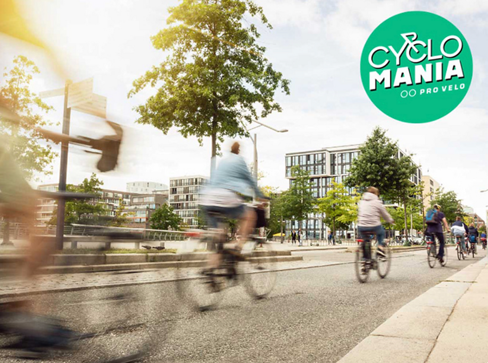People cycling on an urban street with buildings and trees in the background. Green sign reads "Cyclo Mania." Motion blur conveys speed.