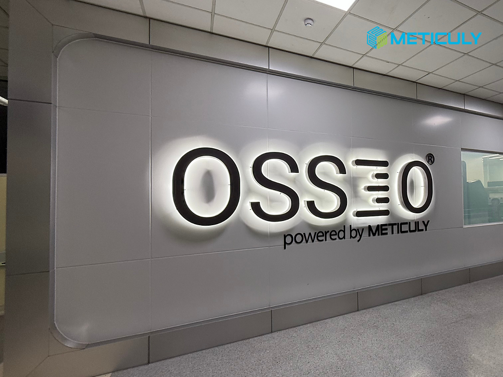 Factory-in-a-Box" point-of-care manufacturing system at Siriraj hospital with a wall-mounted sign with the word 'OSS3O' in illuminated lettering, with 'powered by METICULY' below it.