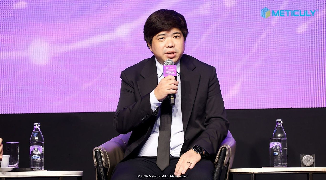 Medium close-up of Meticuly co-founder, Asst. Prof. Dr. Chedtha Puncreobutr, speaking into a microphone during a panel session against a purple abstract digital stage background.