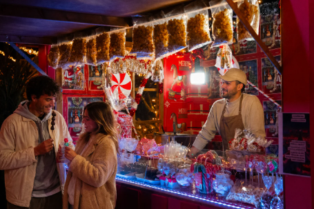 Couple chats at a festive candy stall as a smiling vendor stands behind jars of sweets, with red holiday decor and lights.