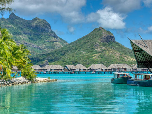 Overwater bungalows in Tahiti surrounded by turquoise lagoon waters, with lush green mountains rising in the background under a clear sky.