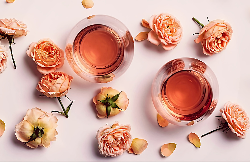 Two glasses of Rose Wine surrounded by pink roses and rose petals on a pink background.