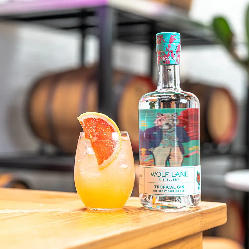Cocktail with an orange slice next to a bottle of Wolf Lane Tropical Gin on a wooden bar. Barrels and plants blurred in the background.