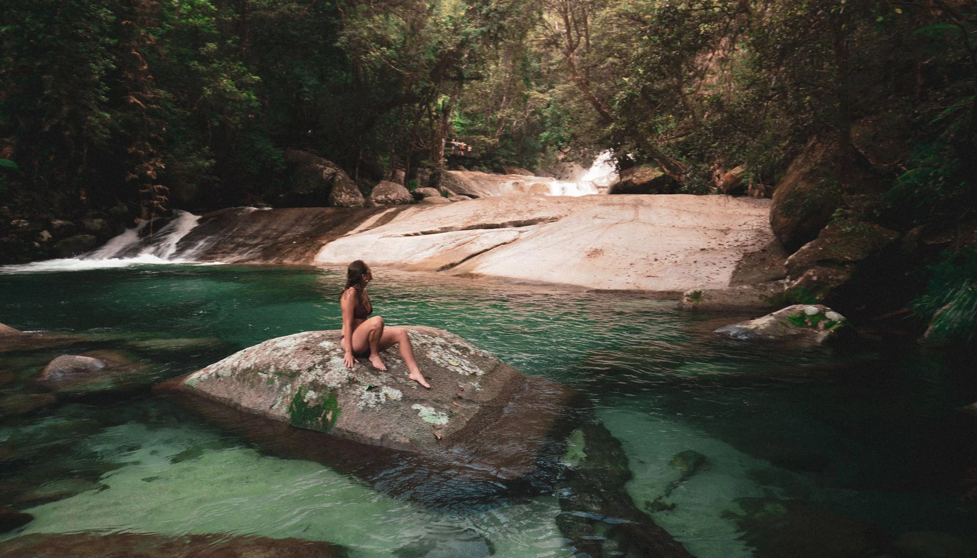 A woman in a swimsuit sits on a rock in a peaceful forest river, surrounded by lush greenery and clear water. Serenity and nature abound.