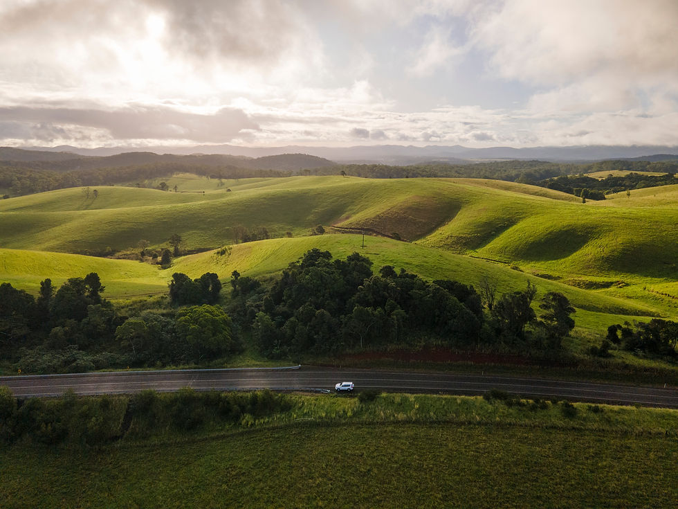 A white car drives along a road through lush green hills with trees, under a cloudy sky. The scene is serene and expansive.