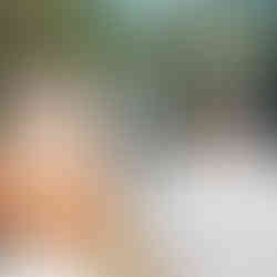 Two people stand outdoors facing a waterfall surrounded by lush green trees. The woman's hair is long and wavy. Bright and serene scene.