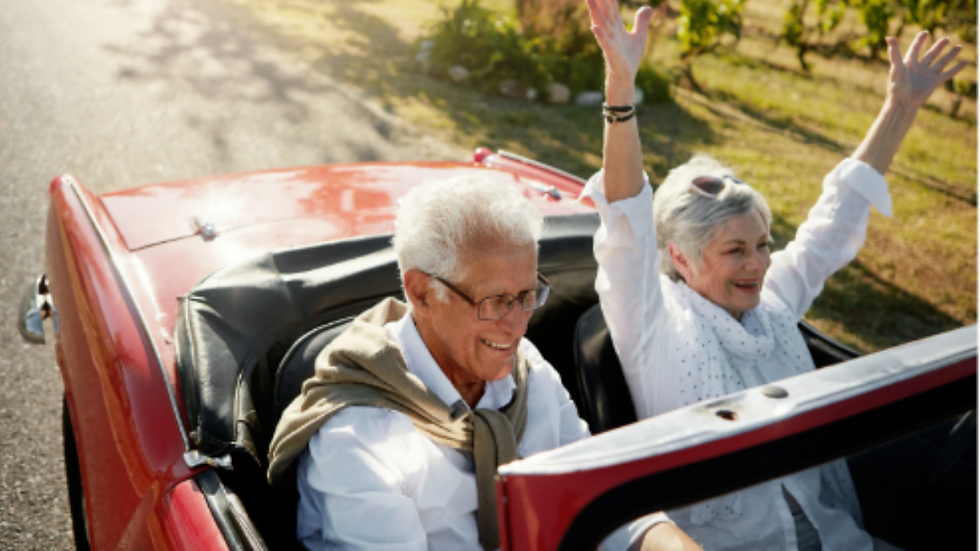 Elderly couple joyful in red convertible, woman raises arms in excitement. Sunny rural road, positive mood.