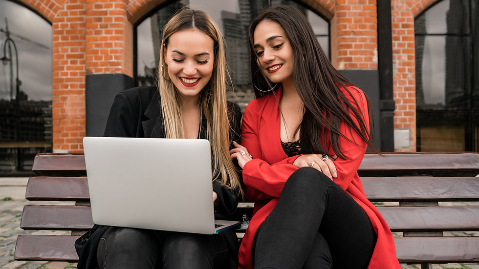 Two women sit on a bench, smiling while looking at a laptop. They wear red and black outfits. Background: red brick building.