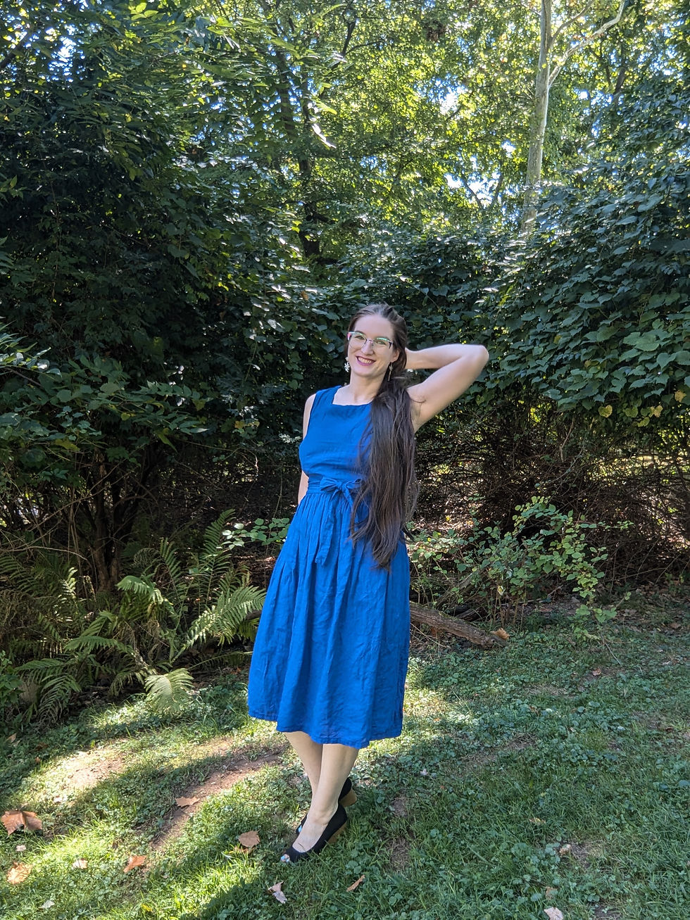A white woman with long brown hair and glasses wearing a royal apron blue dress.