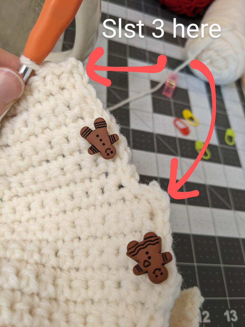 One side of one flap of the tea cozy is shown - the side where the handle will go. Two buttons shaped like gingerbread people have been added - one in the space that will wrap around the space underneath the handle and another to the space that will go immediately under the handle. Text "slip stitch 3 here" has arrows pointing to the outer corners along the sides of the flaps.