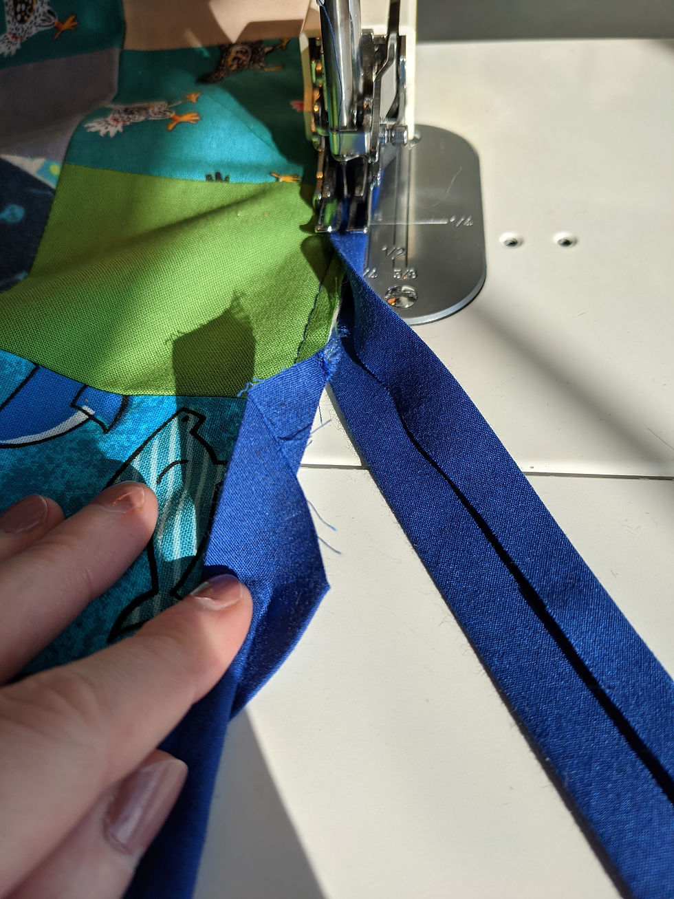 A quilt edge being bound with bias tape by a sewing machine. The blue cotton bias tape is wrapped around the edges of the quilt. The beginning of the strip of bias tape is raw, the other end of the bias tape approaches the beginning.