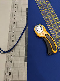 Blue cotton fabric folded into the double triangle as described. The side with two folds has been cut using a rotary cutter so that a very narrow strip of fabric is coming off of that side of the triangle. The rotary cutter and a quilter's rule sit on top of the fabric.