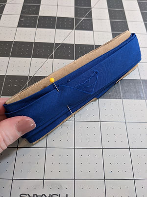 A cardboard rectangle with a length of blue cotton bias tape wrapped around it. The end of the bias tape is held in place with a sewing pin.