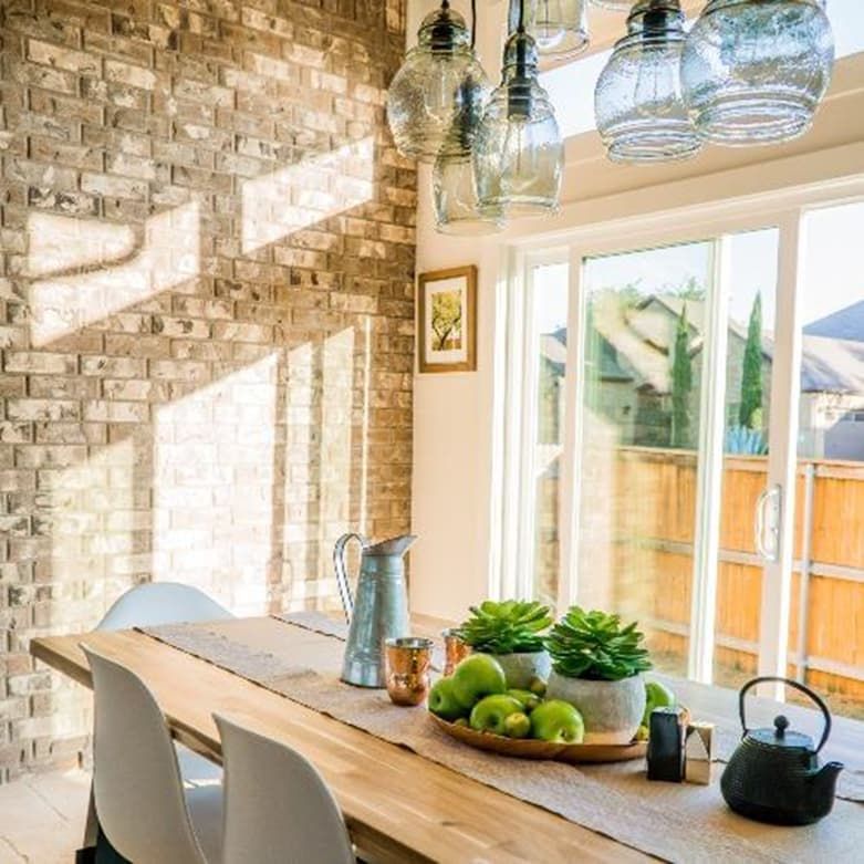 Een goed verlichte woonkamer, veel natuurljik licht van buiten. De woonkamer heeft stenen muren en er staat een tafel met planten er op. Ook hangen er lampen.