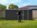A man holding a drill and a phone in front of a large garden shed.