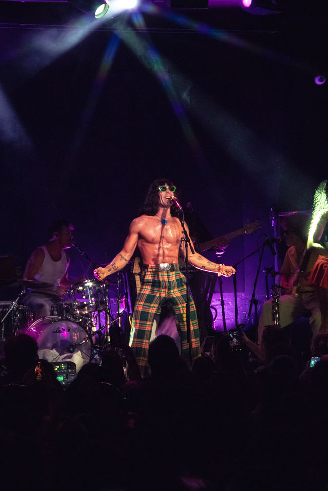 Shirtless performer singing on stage with band.