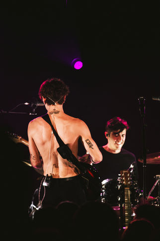 Two musicians performing on stage, one shirtless with guitar.