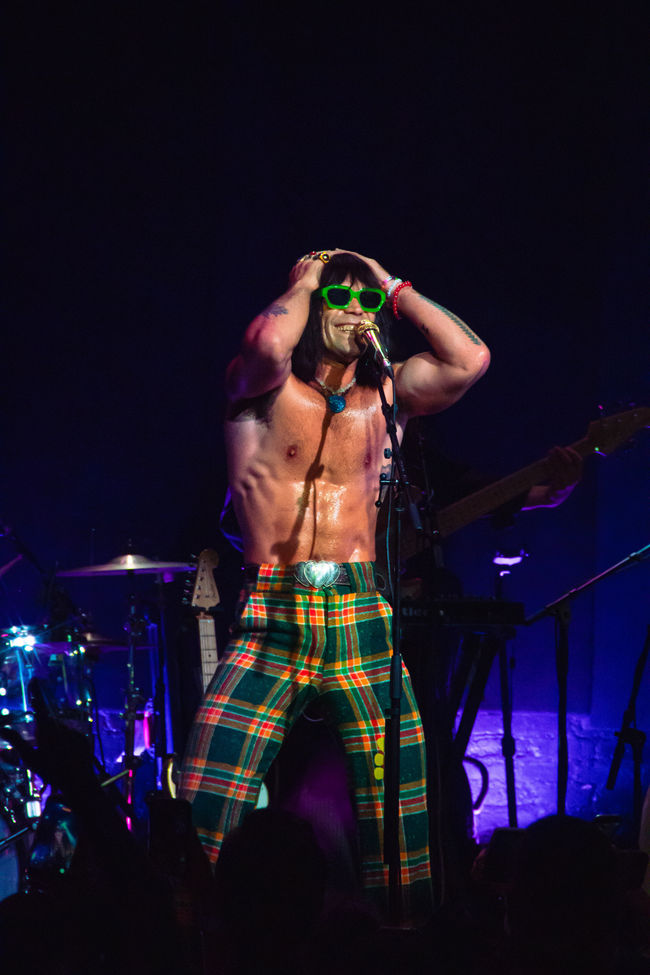 Shirtless performer in plaid pants and green sunglasses.