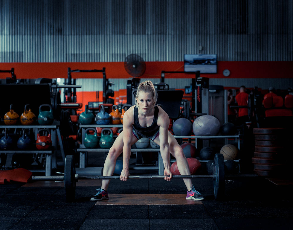 Femme soulevant une barre dans une salle de gym remplie de kettlebells colorés. Ambiance concentrée et déterminée. Mur orange et gris.