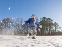 A child in a blue jacket joyfully runs through snow, kicking up powder. Clear sky and trees in the background on a sunny winter day.