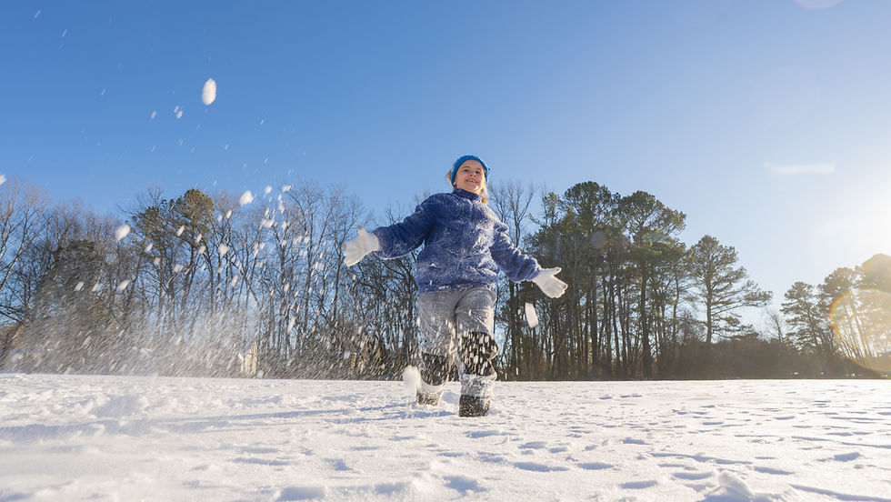 A child in a blue jacket joyfully runs through snow, kicking up powder. Clear sky and trees in the background on a sunny winter day.