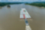 Aerial view of a towboat pushing a large barge of multicolored containers on a wide river, flanked by lush green forests under a cloudy sky.