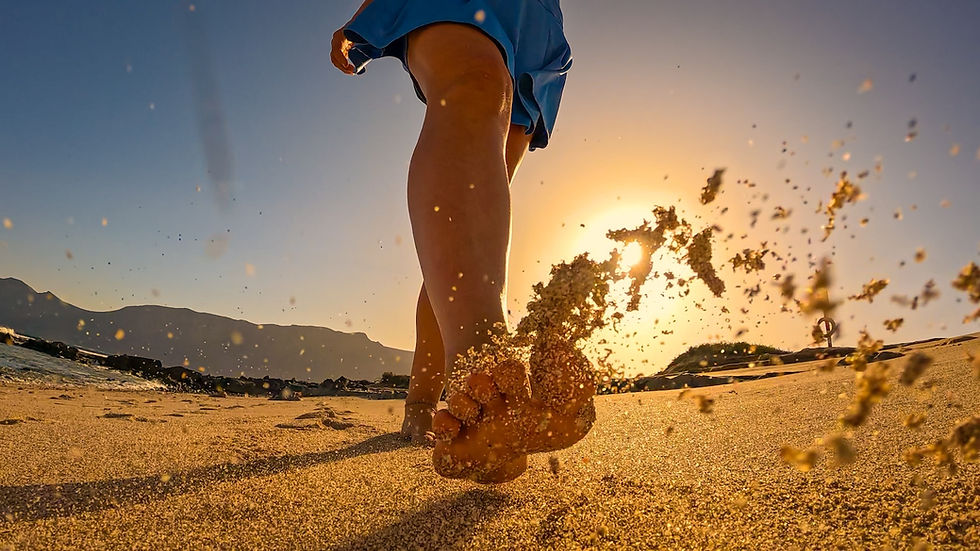 A low-angle view of a person in blue shorts kicking sand on a beach at sunset, with golden light and mountains in the background.