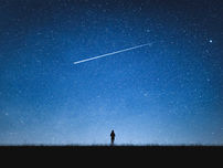 A silhouette of a person stargazing under a vast, starry sky with a bright meteor trail.