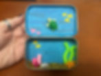 A hand holding an open tin with aquatic diorama: green turtle, colorful fish, beads, and shells on blue background, resembling a mini ocean scene.