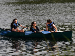Three people in a canoe paddle on a calm lake. They're wearing blue life vests and chatting, with rippled water in the background.