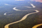 A meandering brown river winds through dense green forest, visible from an aerial view, with fields in the distant background.