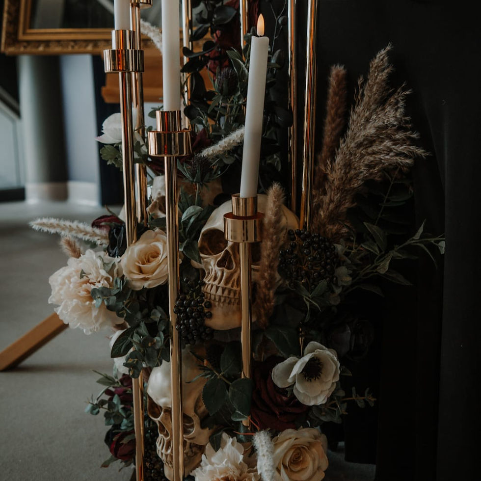 Candles in gold holders with skulls, roses, and pampas grass. Dark, moody setting with a black backdrop.