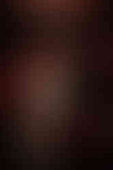 couple stood in a dark bar, with neon lights. one is wearing a white short dress, with socks and vans, holding a red bouquet and has long curly blonde hair, the other hair brown long hair and a beard, and is wearing a suit jacket, band tshirt and black trousers. both are smiling