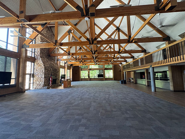 Large empty Camp Tracy lodge hall with stone fireplace, exposed timber rafters, tall windows, and an upper balcony.