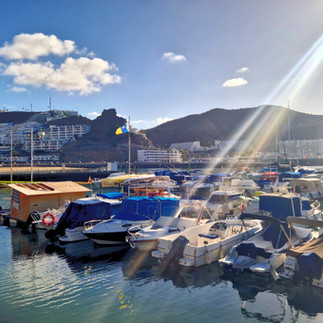 Sunlit marina in Puerto Rico, Gran Canaria, with small boats, mountains, and reflections on the water.