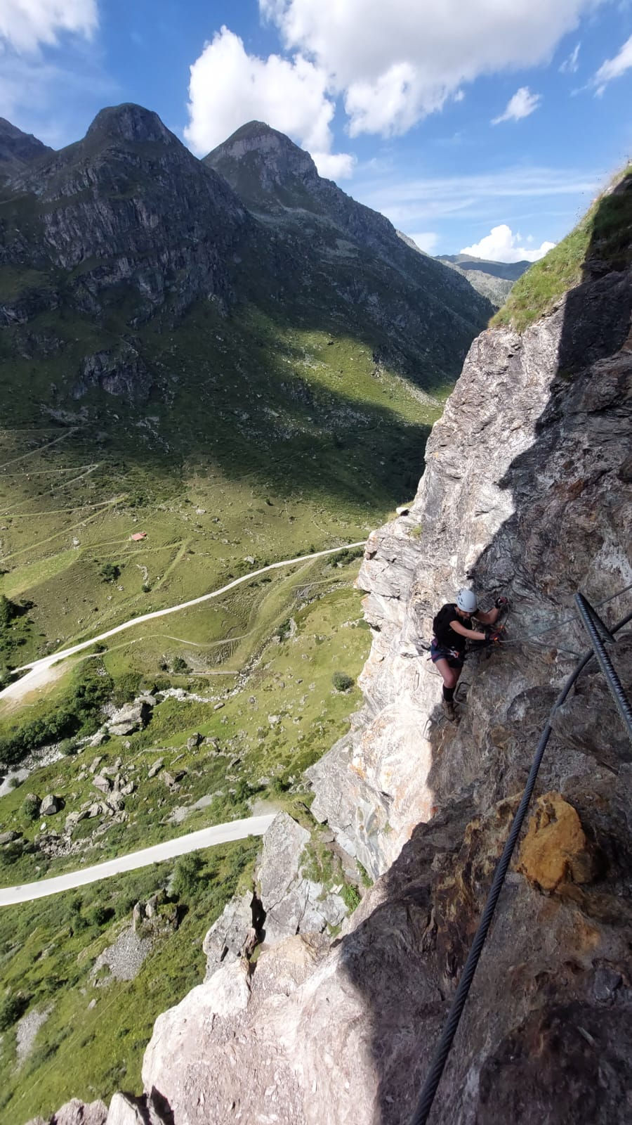 Climber ascends a rocky cliff with safety gear in a mountainous landscape. Blue sky, white clouds, and verdant terrain in the background.