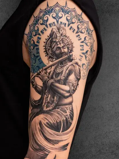 A devotional black-and-grey tattoo of Lord Krishna playing the flute, placed on the upper arm. This piece embodies peace and divinity with intricate detailing and a radiant aura around Krishna’s head. Created by students of Aliens School of Tattoo Art
