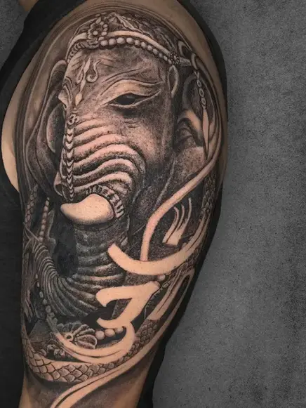 An intricate Lord Ganesha tattoo with traditional elements, located on a male's upper arm, done in realistic black and grey shading.