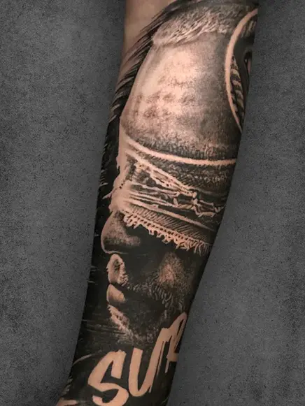 A tattoo of a blindfolded man's face with dramatic shadows, stretching along a male's forearm, in a moody black and grey realism style.