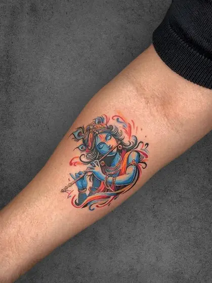 A vibrant tattoo of Lord Krishna in blue and red tones, playing a flute with flowing accents, placed on the forearm