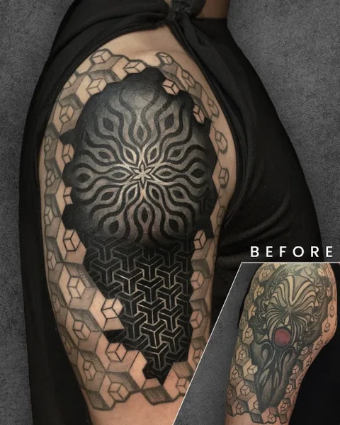 A mesmerizing geometric design cover-up featuring a dark, intricate pattern with layers of symmetry. The original tattoo, visible in the corner, is an abstract design with a red gem at its center