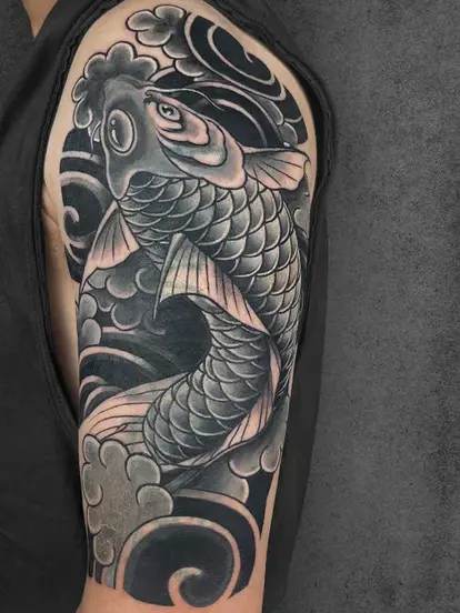 A detailed black-and-grey tattoo of a koi fish flowing through dark waters with swirling waves, placed on the upper arm. The design symbolizes perseverance and strength. Created by students of Aliens School of Tattoo Art.