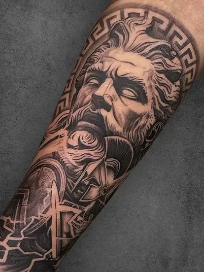 A detailed arm tattoo featuring Zeus, the Greek god of thunder, with lightning bolts and a decorative Greek key pattern framing his majestic visage.